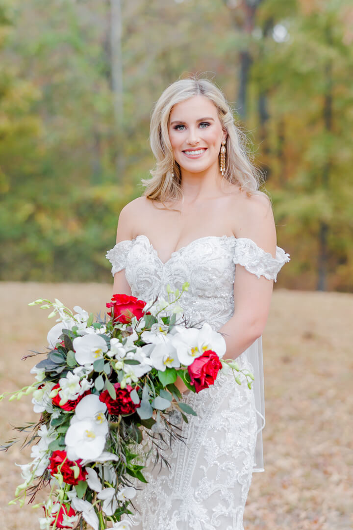 A bride in a white lace wedding gown stands in a field holding her large bouquet with red roses grandeur house wedding