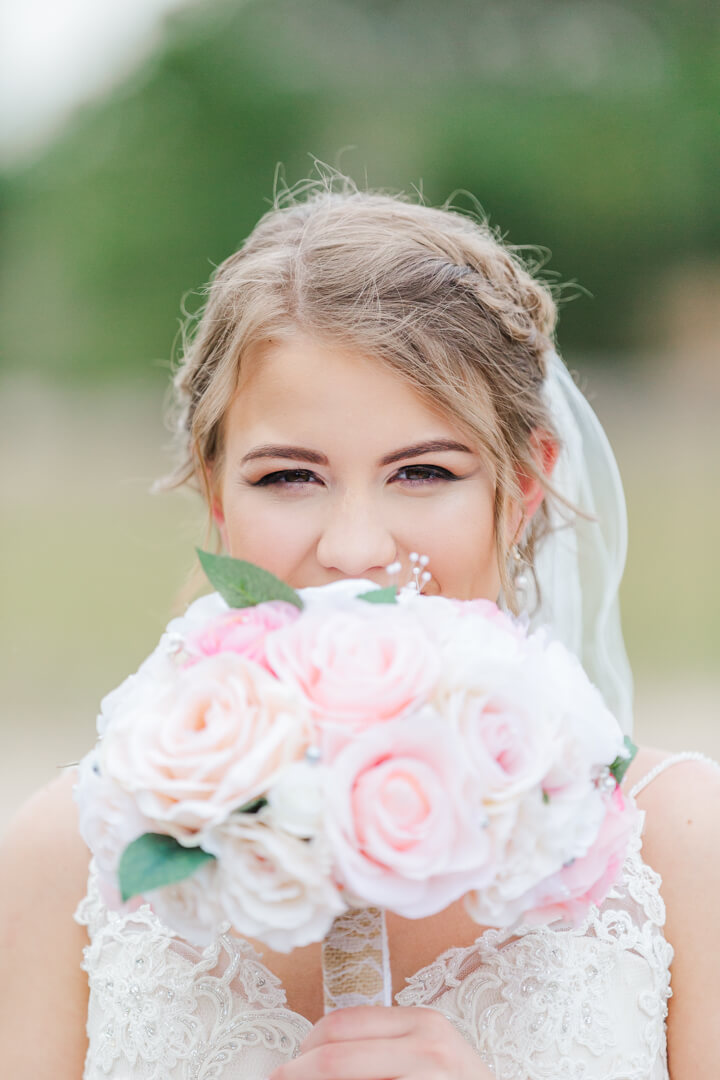 A bride in a white lace dress hides her smile behind her pink bouquet grandeur house wedding