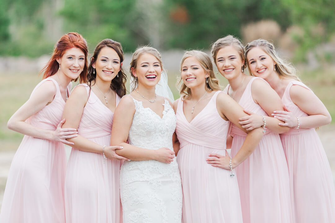 A bride stands in a grassy field in a white lace wedding gown with her bridesmaids wearing pink heritage acres wedding
